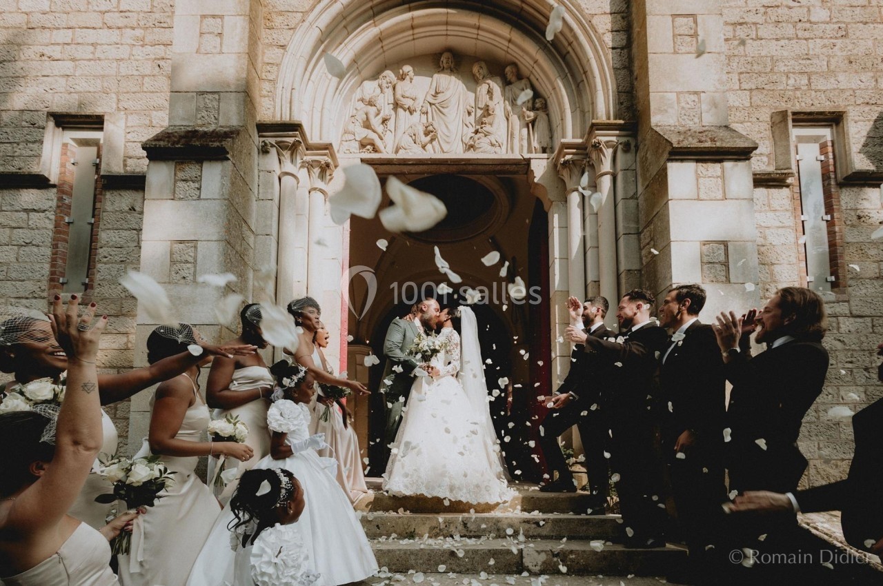 Un couple de mariés sort d'une église sous une pluie de pétales, entouré d'invités applaudissant.