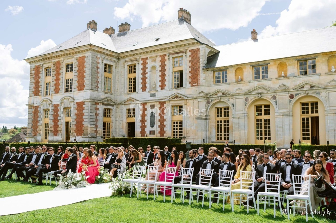 Invités assis en plein air devant un château lors d'une cérémonie, sous un ciel partiellement nuageux.