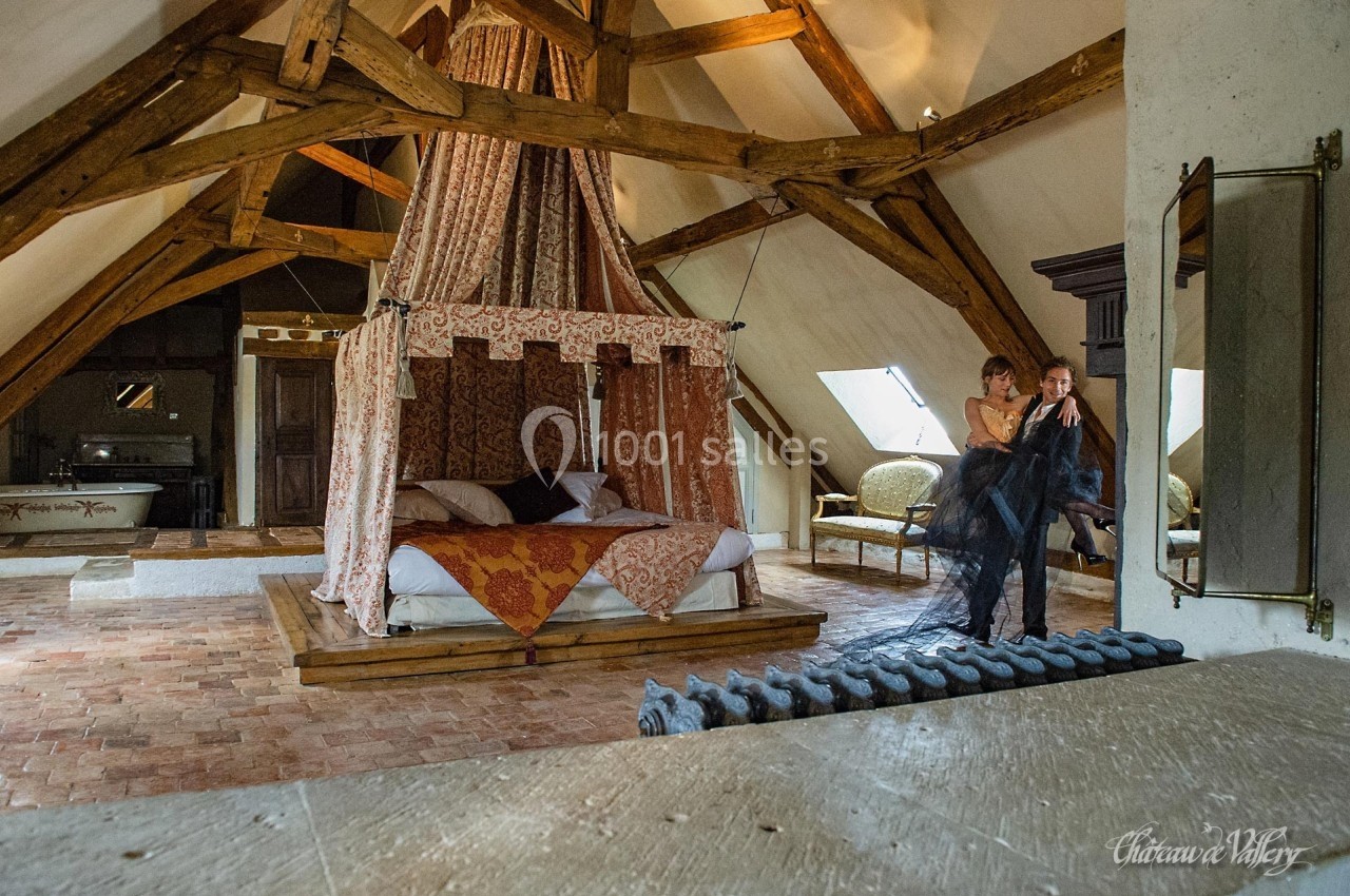 Chambre mansardée avec lit à baldaquin, poutres apparentes et un couple debout près d'une fenêtre.