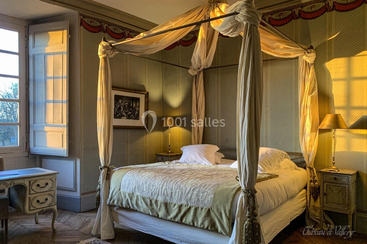 Lit à baldaquin dans une chambre élégante avec murs tapissés, lumière naturelle et mobilier ancien.