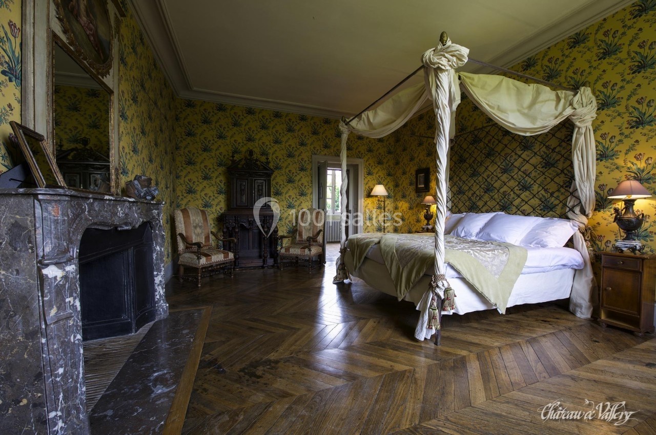 Chambre avec lit à baldaquin, parquet en bois, cheminée en marbre et murs ornés de motifs floraux jaunes.