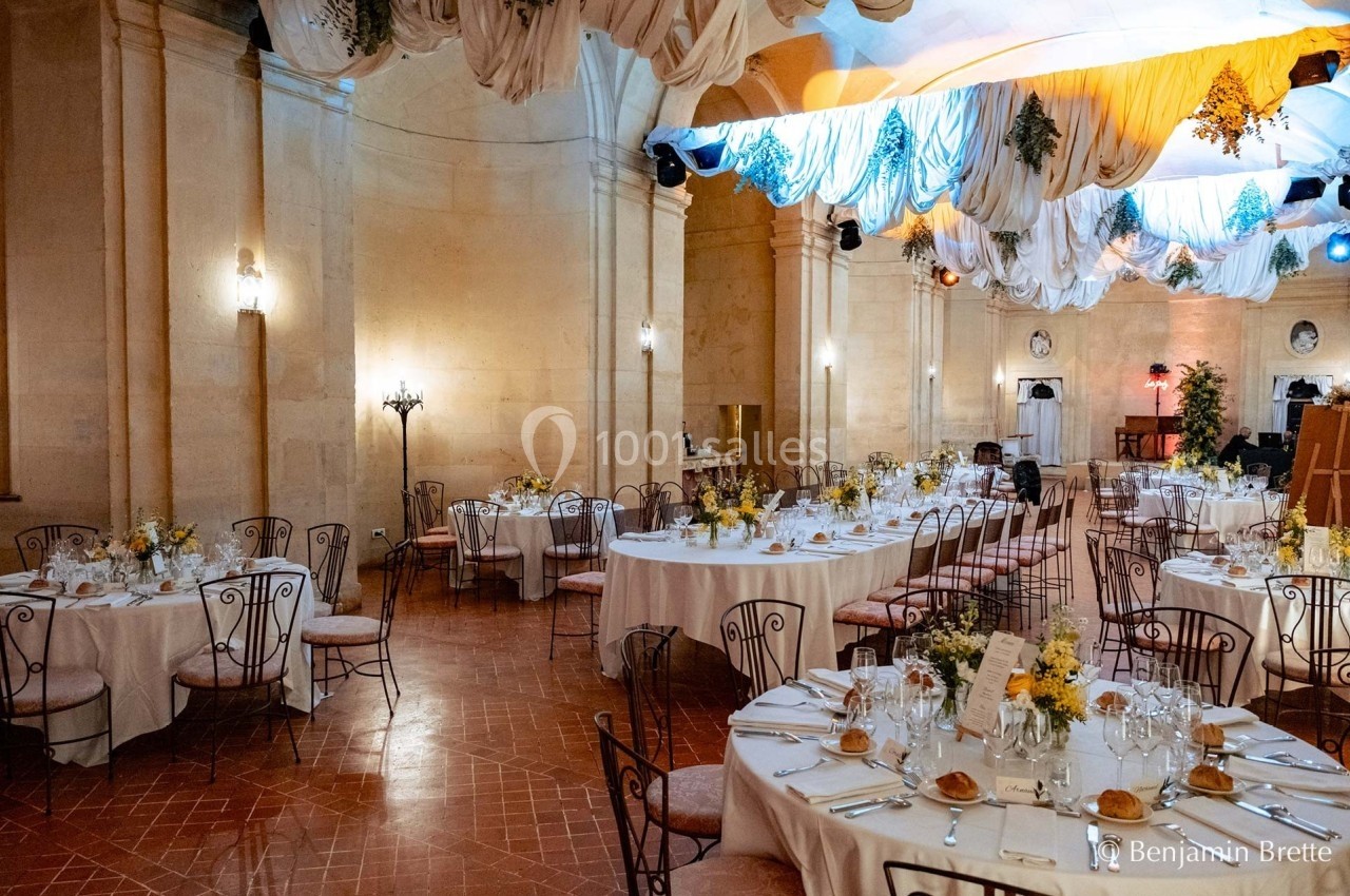 Salle de réception décorée avec des tables rondes dressées, des chaises en fer forgé et un plafond orné de tissus suspendus.