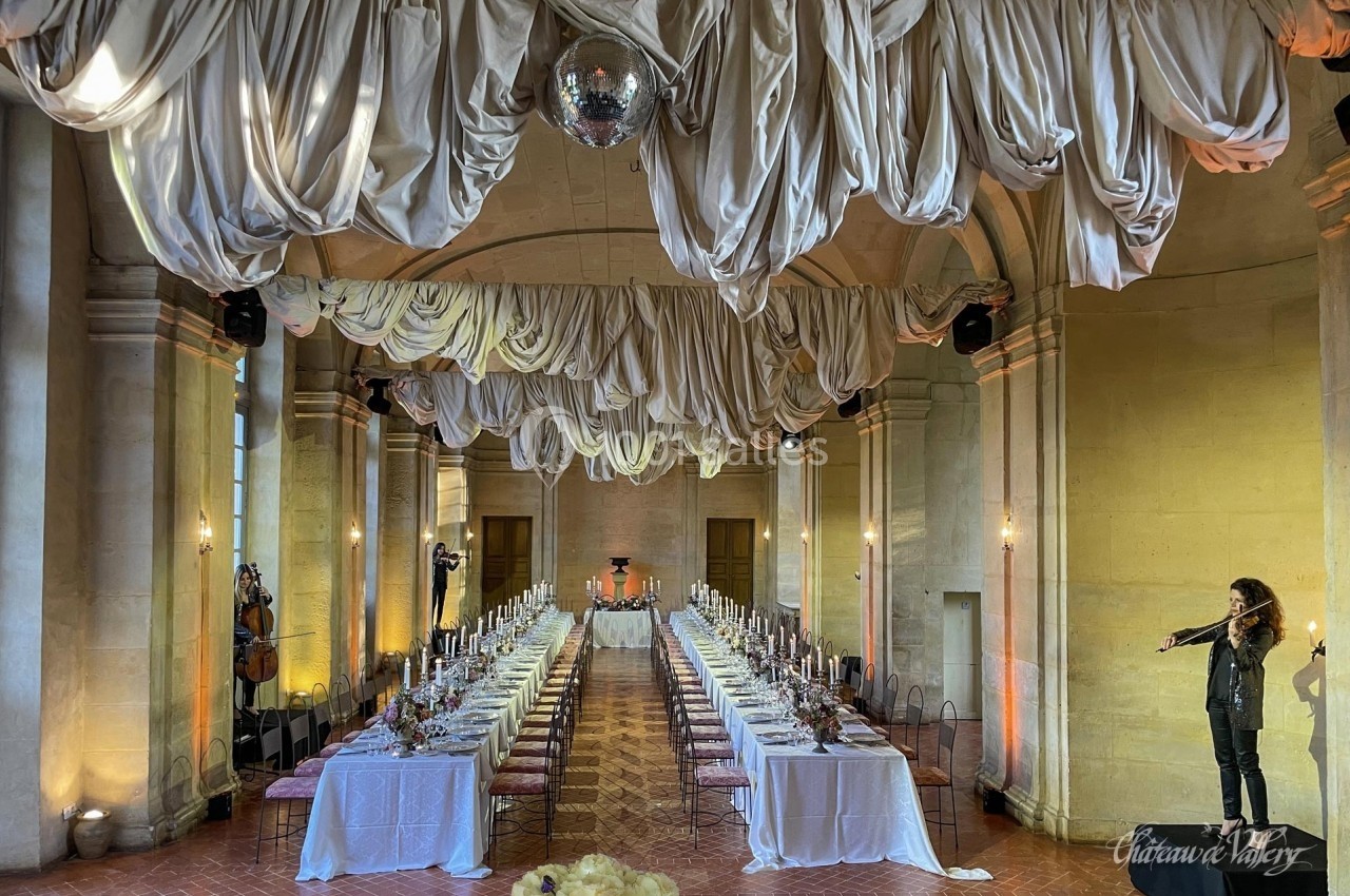 Grande salle avec tables dressées pour un banquet, plafond décoré de drapés et musicienne jouant du violon à droite.