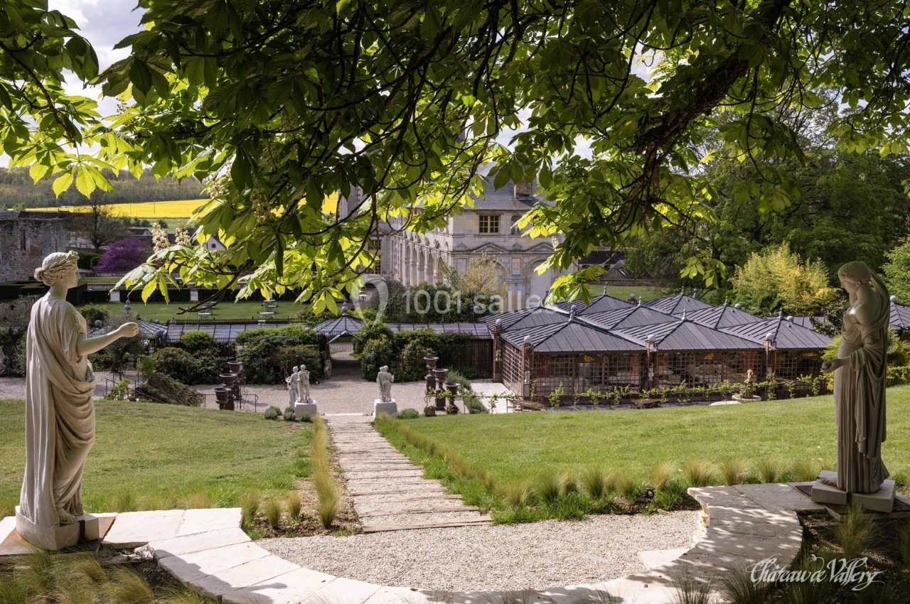 Allée bordée de statues menant à un jardin avec pavillons, sous un arbre aux larges feuilles.