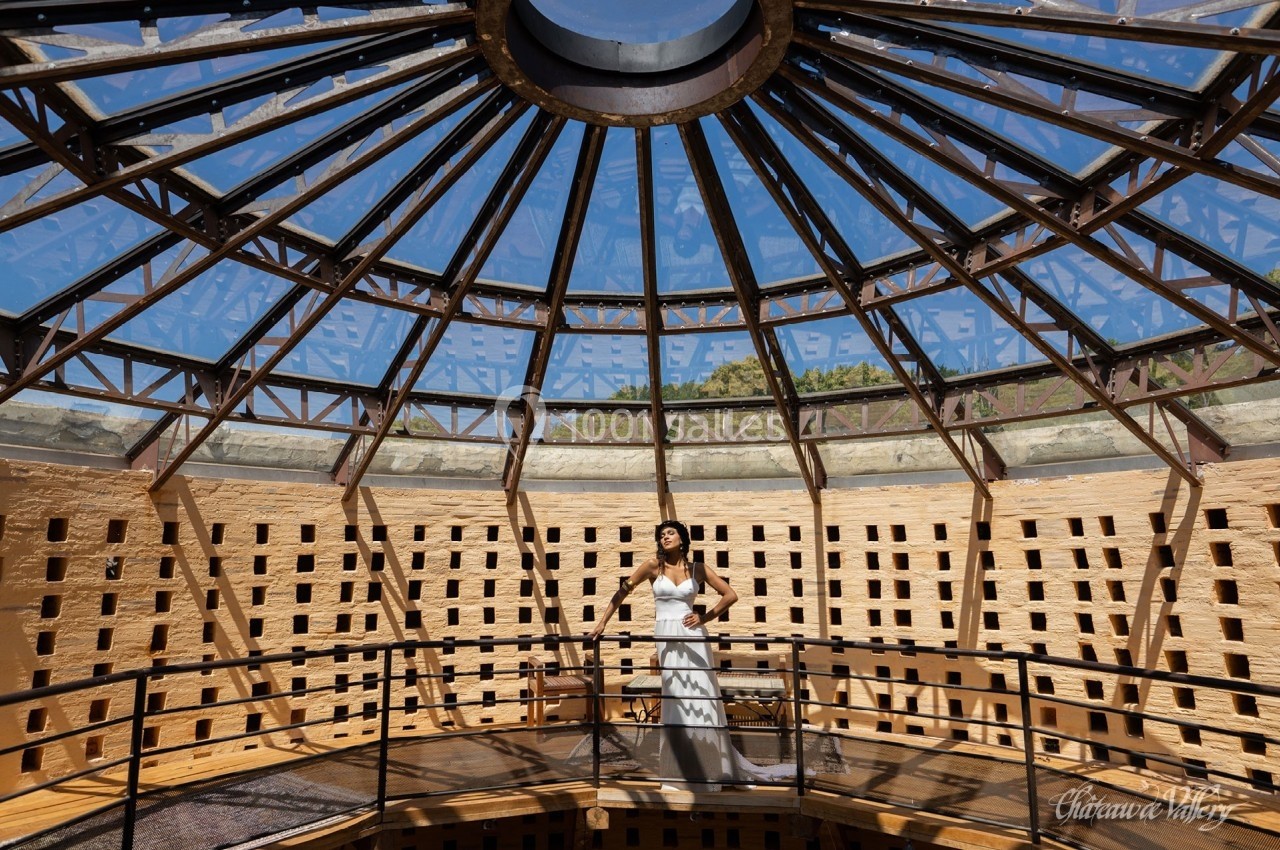 Une femme en robe blanche se tient sur une passerelle sous une verrière circulaire en métal et verre.