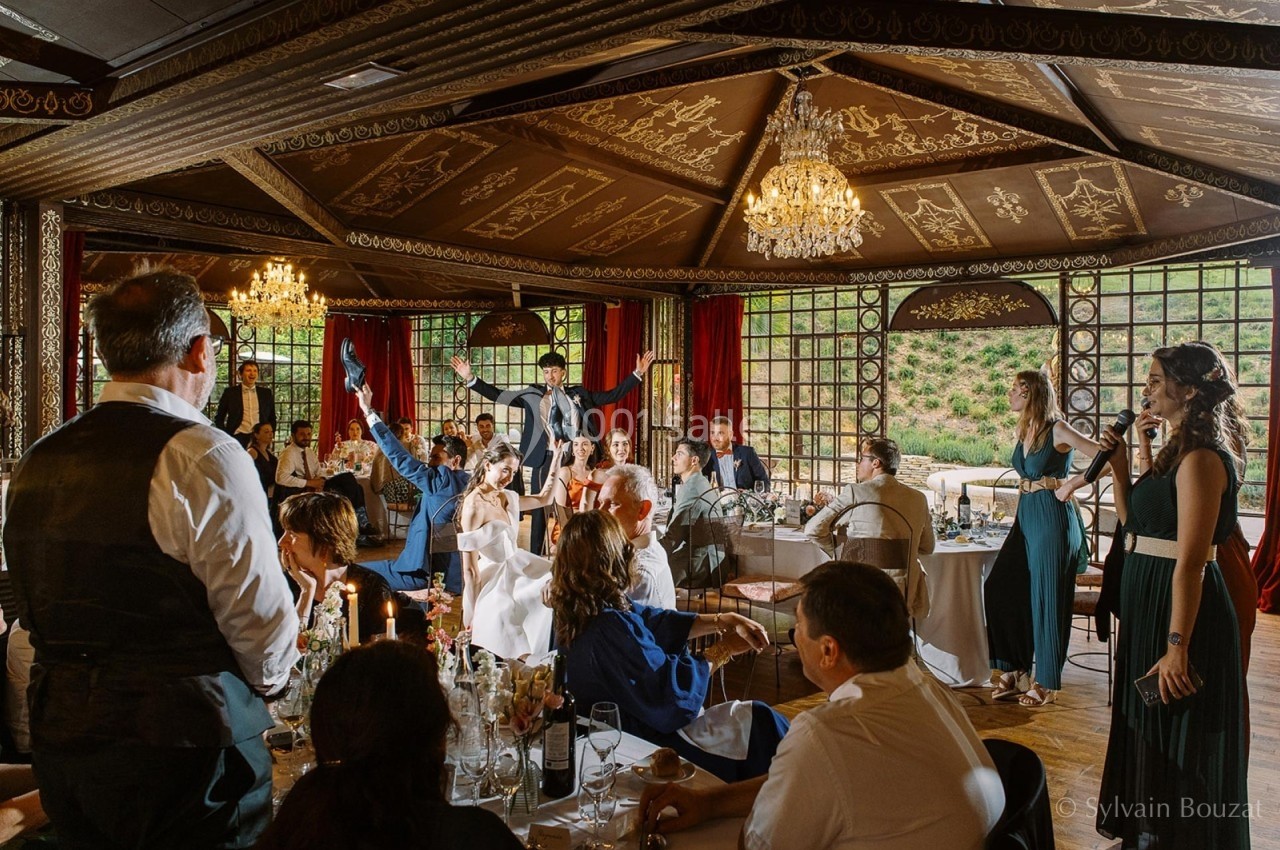 Des convives assis et debout dans une salle décorée, participant à une réception festive avec des chandeliers et des rideaux…