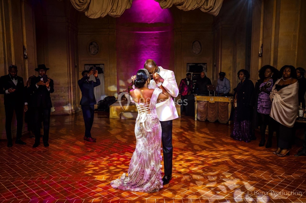 Un couple danse au centre d'une salle élégante, éclairée par des lumières colorées, entouré d'invités attentifs.
