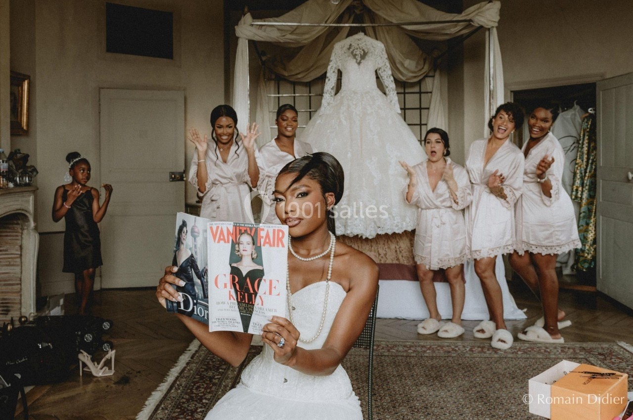 Une mariée en robe blanche lit un magazine, entourée de demoiselles d'honneur applaudissant dans une chambre.