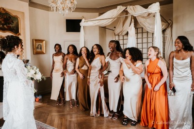 Un groupe de femmes en robes élégantes réagit avec joie face à une mariée dans une pièce décorée.