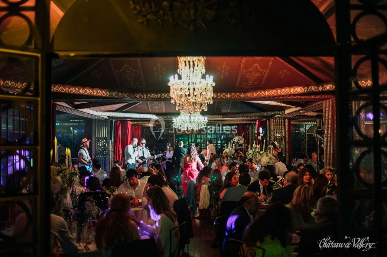 Salle élégante avec des invités dînant sous des lustres, un groupe de musiciens jouant en arrière-plan.