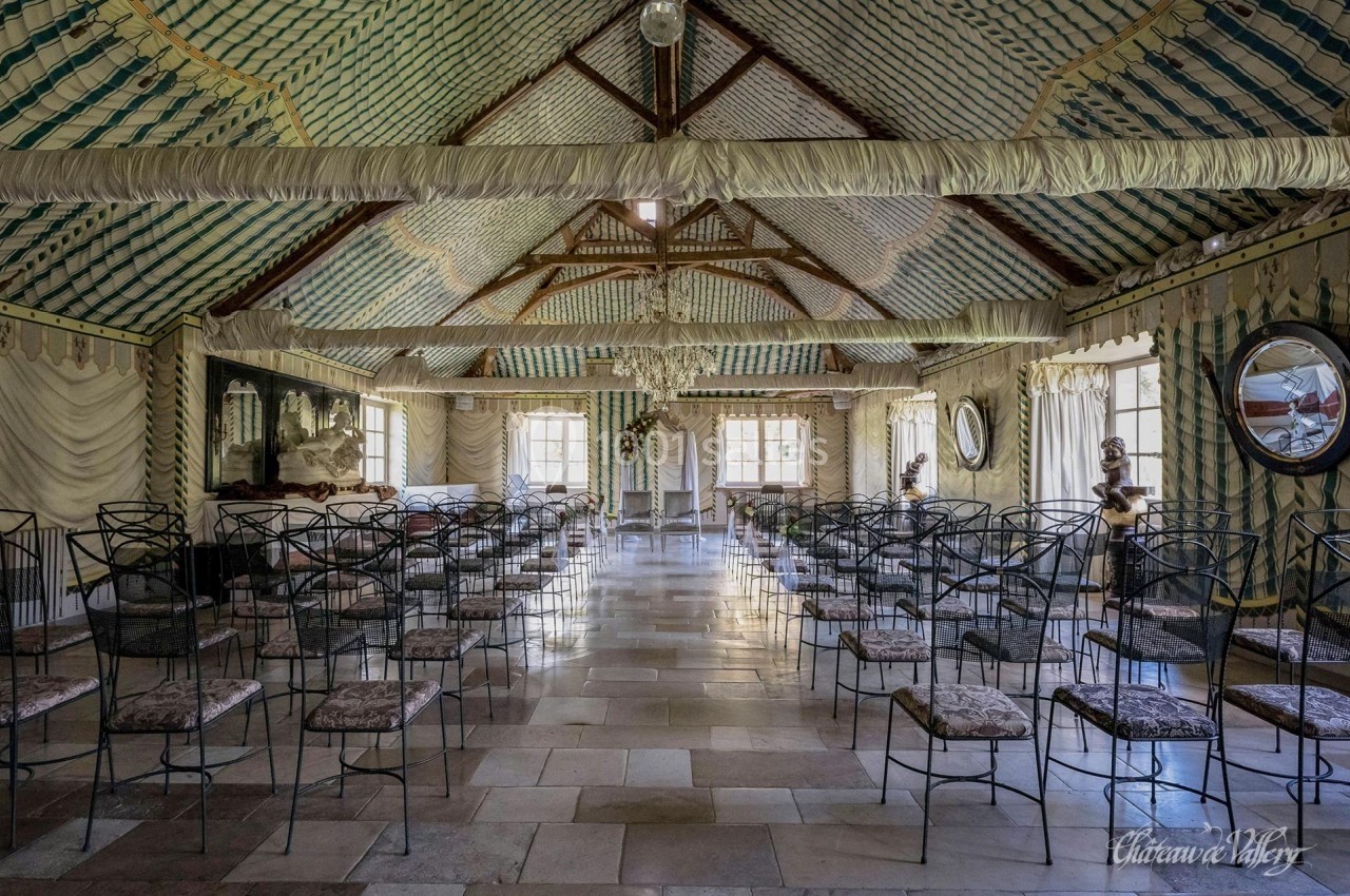 Salle décorée avec des chaises alignées, plafond en tissu rayé et lustre central, éclairée par des fenêtres.