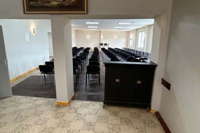 Des personnes, dont des enfants, entrent dans un bâtiment à façade claire avec des fenêtres et une porte ouverte.