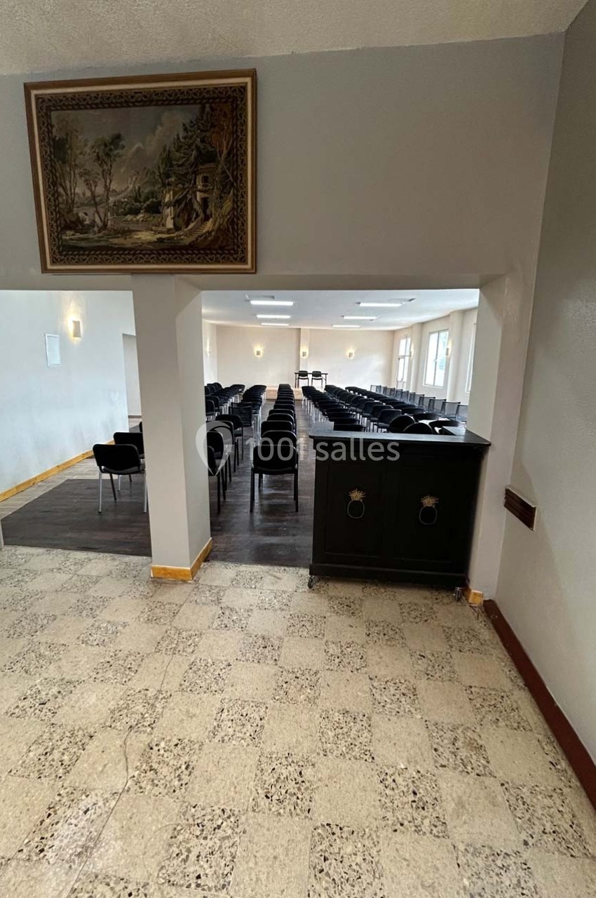 Vue d'une salle de réunion avec rangées de chaises noires alignées, éclairée par des fenêtres et des plafonniers.