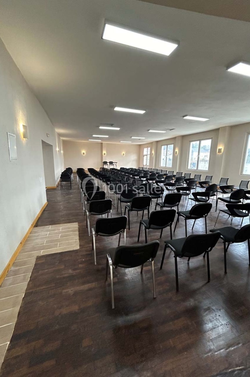 Salle lumineuse avec des rangées de chaises noires alignées sur un sol en bois sombre, éclairée par des plafonniers.