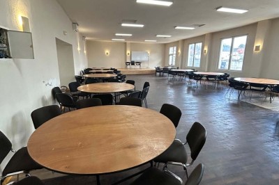 Des personnes, dont des enfants, entrent dans un bâtiment à façade claire avec des fenêtres et une porte ouverte.