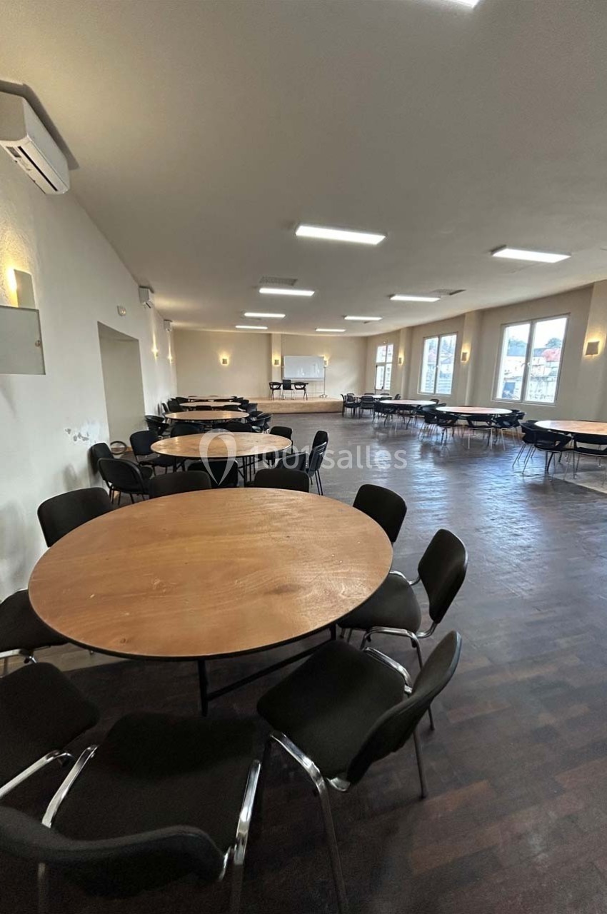 Salle spacieuse avec des tables rondes en bois entourées de chaises noires, éclairée par des fenêtres et des luminaires au…