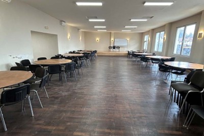 Des personnes, dont des enfants, entrent dans un bâtiment à façade claire avec des fenêtres et une porte ouverte.