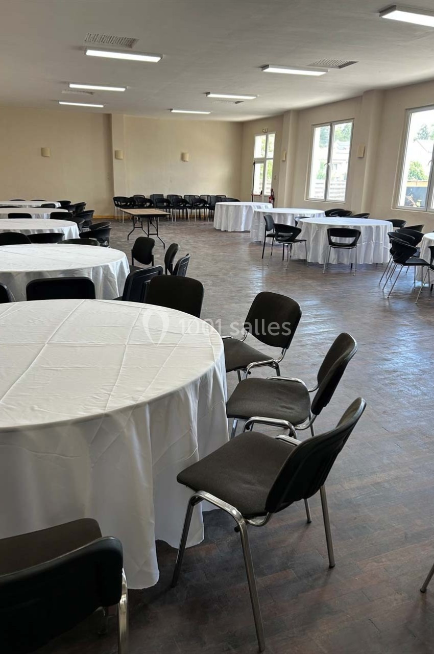 Salle lumineuse avec des tables rondes recouvertes de nappes blanches et des chaises noires disposées autour.