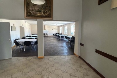 Des personnes, dont des enfants, entrent dans un bâtiment à façade claire avec des fenêtres et une porte ouverte.