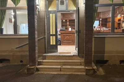 Des personnes, dont des enfants, entrent dans un bâtiment à façade claire avec des fenêtres et une porte ouverte.