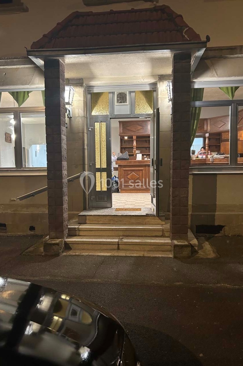 Entrée éclairée d'un bâtiment avec des marches, une porte vitrée et un intérieur visible à travers les fenêtres.