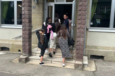 Des personnes, dont des enfants, entrent dans un bâtiment à façade claire avec des fenêtres et une porte ouverte.