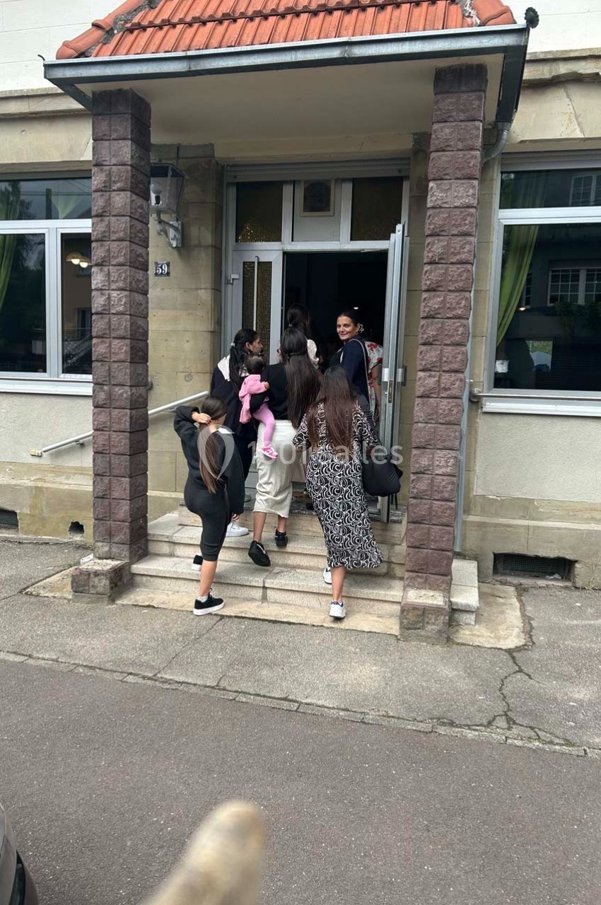 Des personnes, dont des enfants, entrent dans un bâtiment à façade claire avec des fenêtres et une porte ouverte.