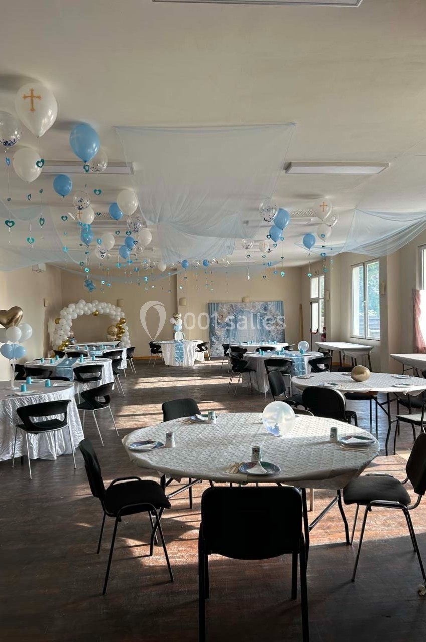 Salle décorée pour une réception avec ballons bleus et blancs, nappes blanches et chaises noires autour des tables.