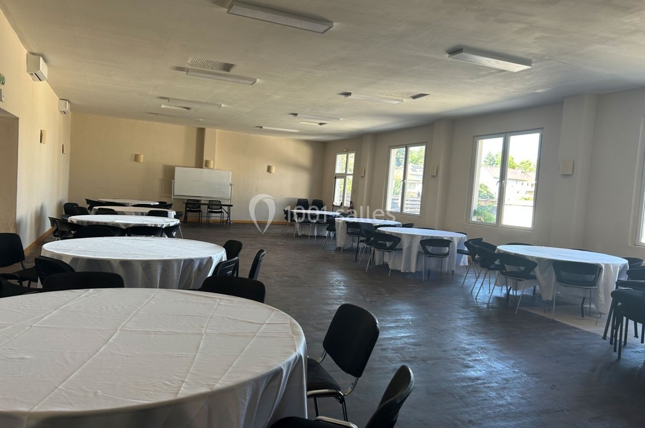 Salle lumineuse avec tables rondes recouvertes de nappes blanches et chaises noires, fenêtres donnant sur l'extérieur.