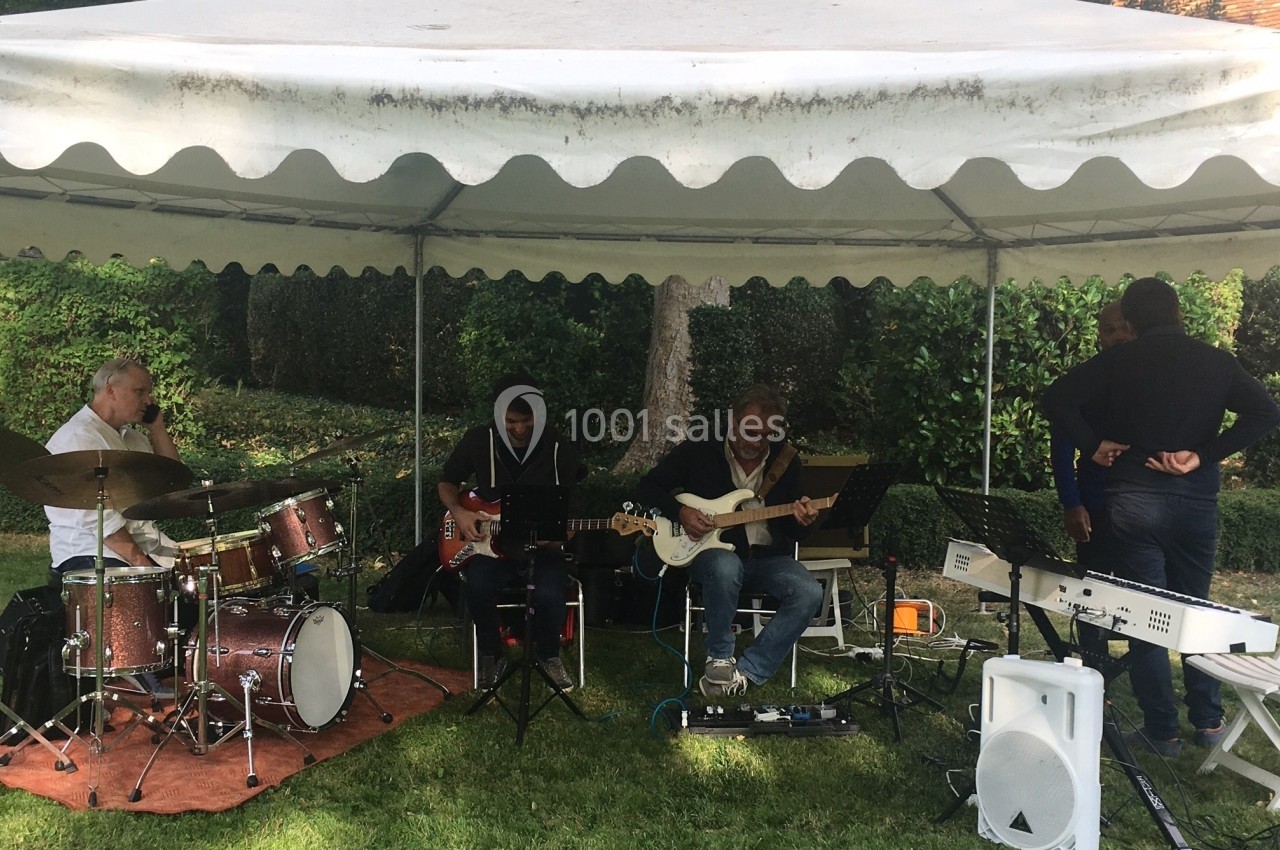 Musiciens jouant sous une tente blanche dans un jardin, avec batterie, guitares et clavier.
