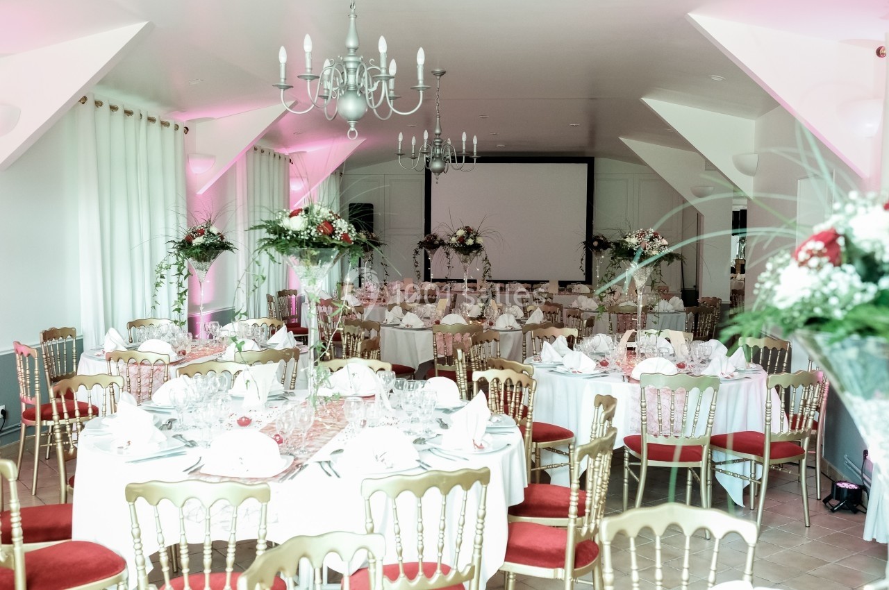 Salle de réception décorée avec des tables rondes, nappes blanches, chaises dorées et arrangements floraux.