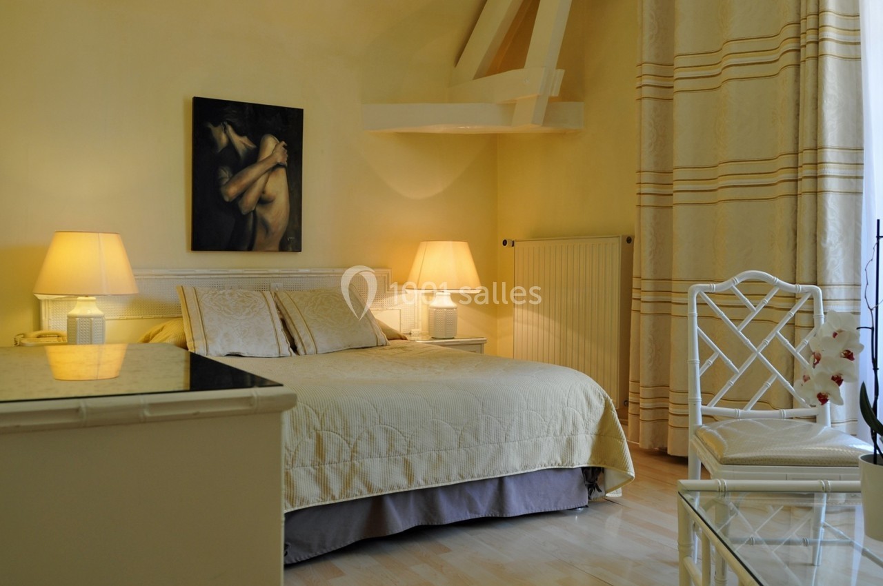 Chambre lumineuse avec lit double, murs jaunes, lampes de chevet, tableau, orchidée et grande fenêtre avec rideaux.