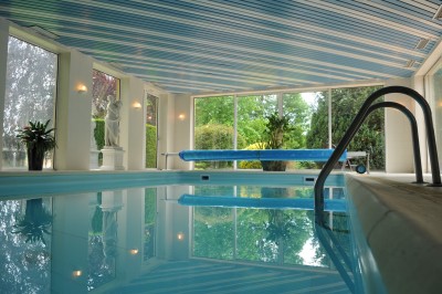 Piscine intérieure avec grandes baies vitrées donnant sur un jardin, entourée de plantes et d'une statue décorative.