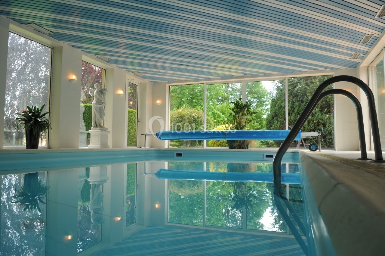 Piscine intérieure avec grandes baies vitrées donnant sur un jardin, entourée de plantes et d'une statue décorative.