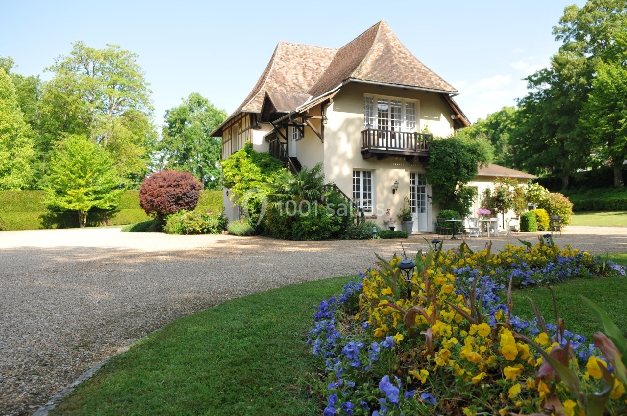 Maison traditionnelle entourée de verdure avec une allée gravillonnée et un parterre de fleurs colorées au premier plan.