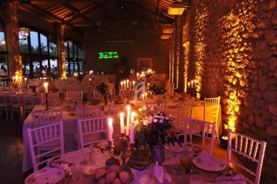 Salle de réception en pierre éclairée par des bougies et des lumières chaudes, avec des tables dressées pour un dîner.