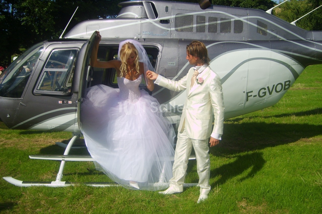 Une mariée en robe blanche descend d'un hélicoptère, aidée par un homme en costume clair, sur une pelouse verdoyante.