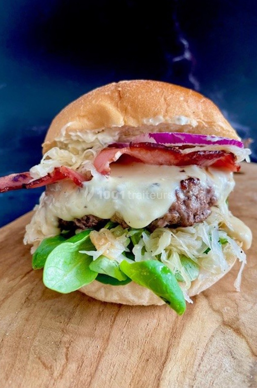 Burger garni de fromage fondu, bacon, oignons rouges, salade verte et choucroute, présenté sur une planche en bois.