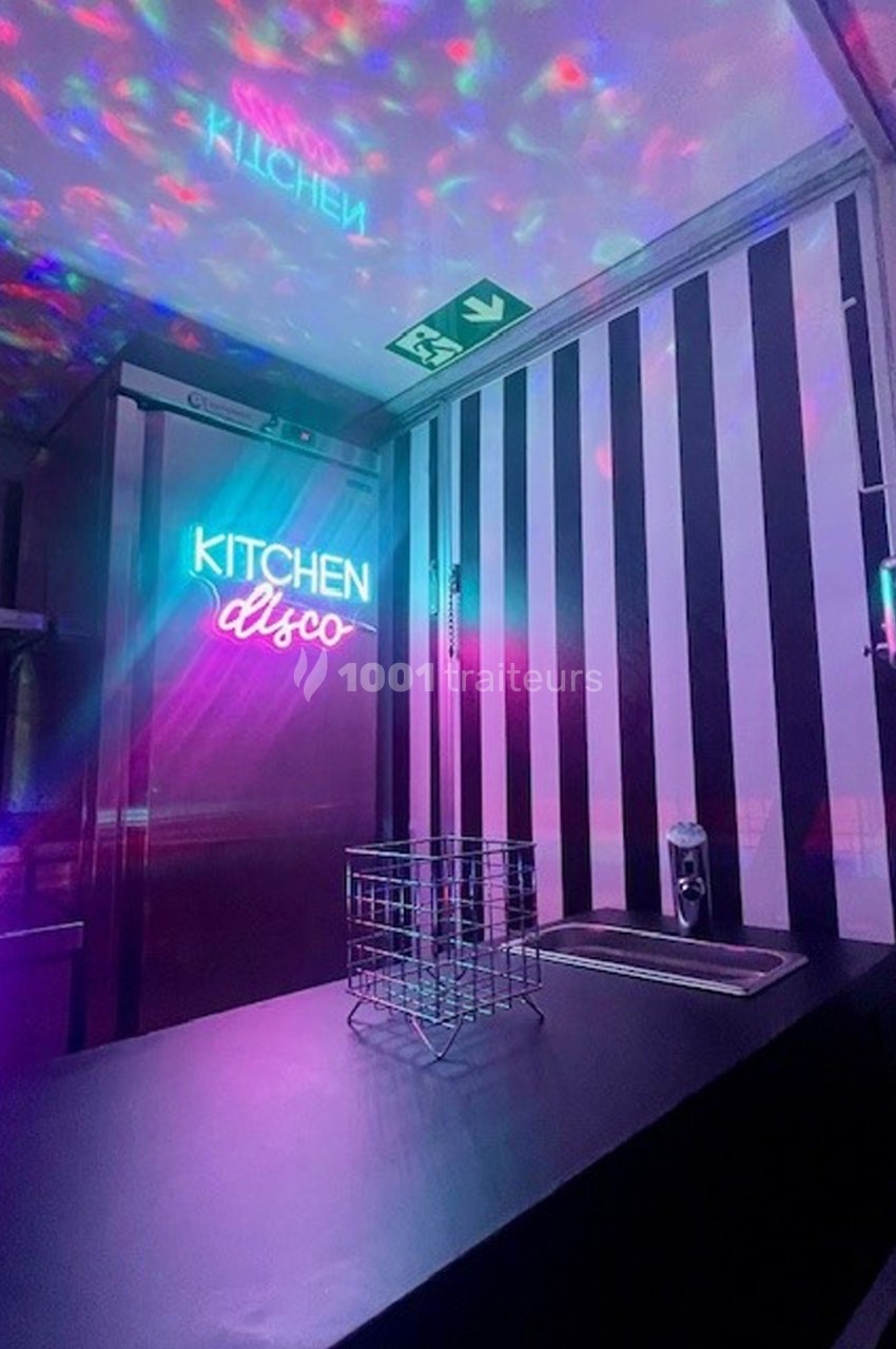 Plan de travail noir avec un évier, mur rayé noir et blanc, enseigne néon ’Kitchen Disco’ et lumières colorées.