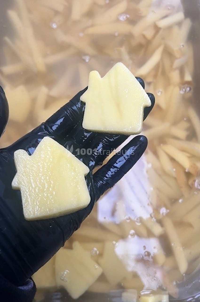 Deux morceaux de pommes de terre découpés en forme de maison, tenus par une main gantée au-dessus d'un bac d'eau.