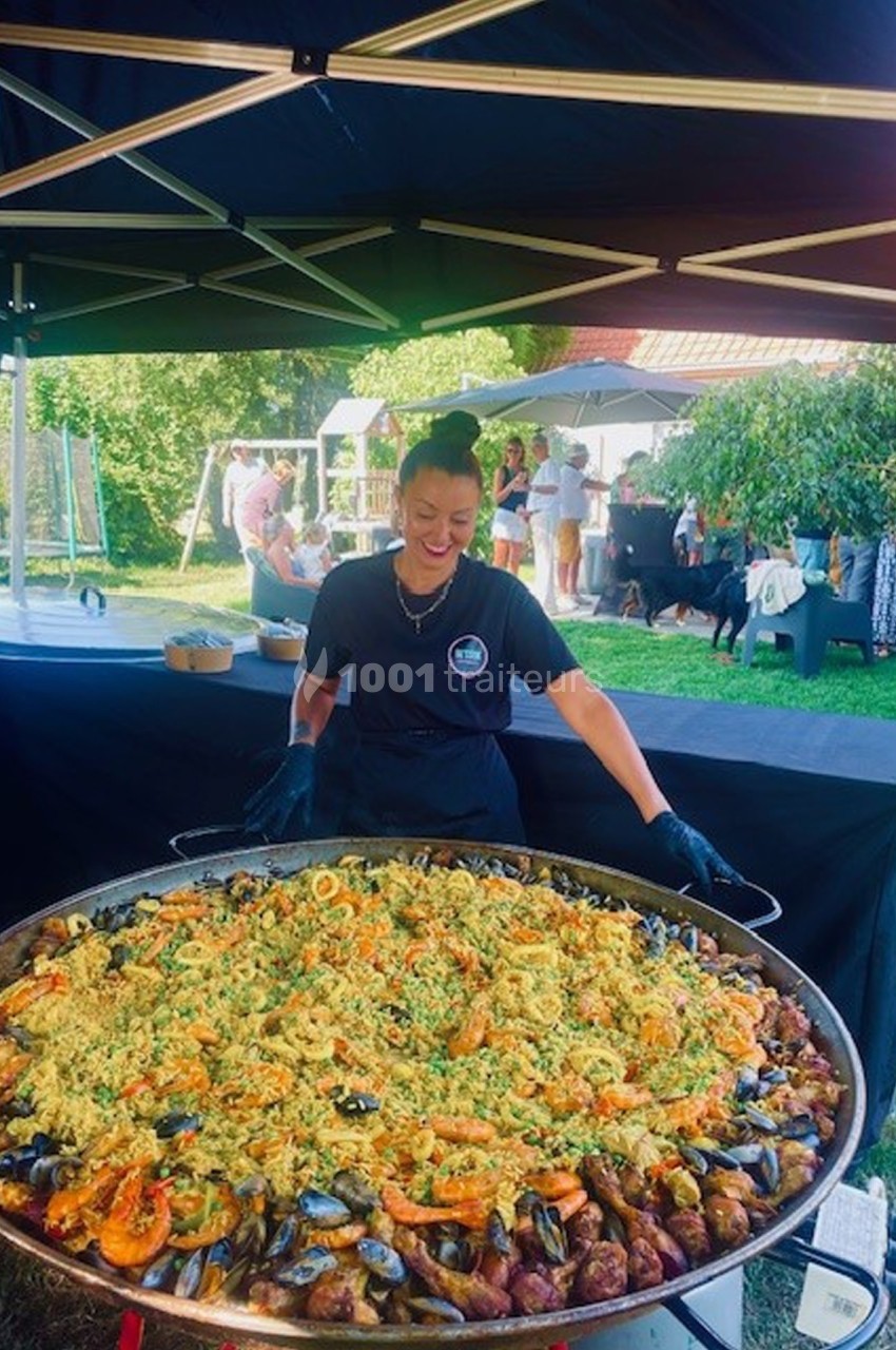 Une femme souriante prépare une grande paella garnie de fruits de mer, sous un stand en extérieur.