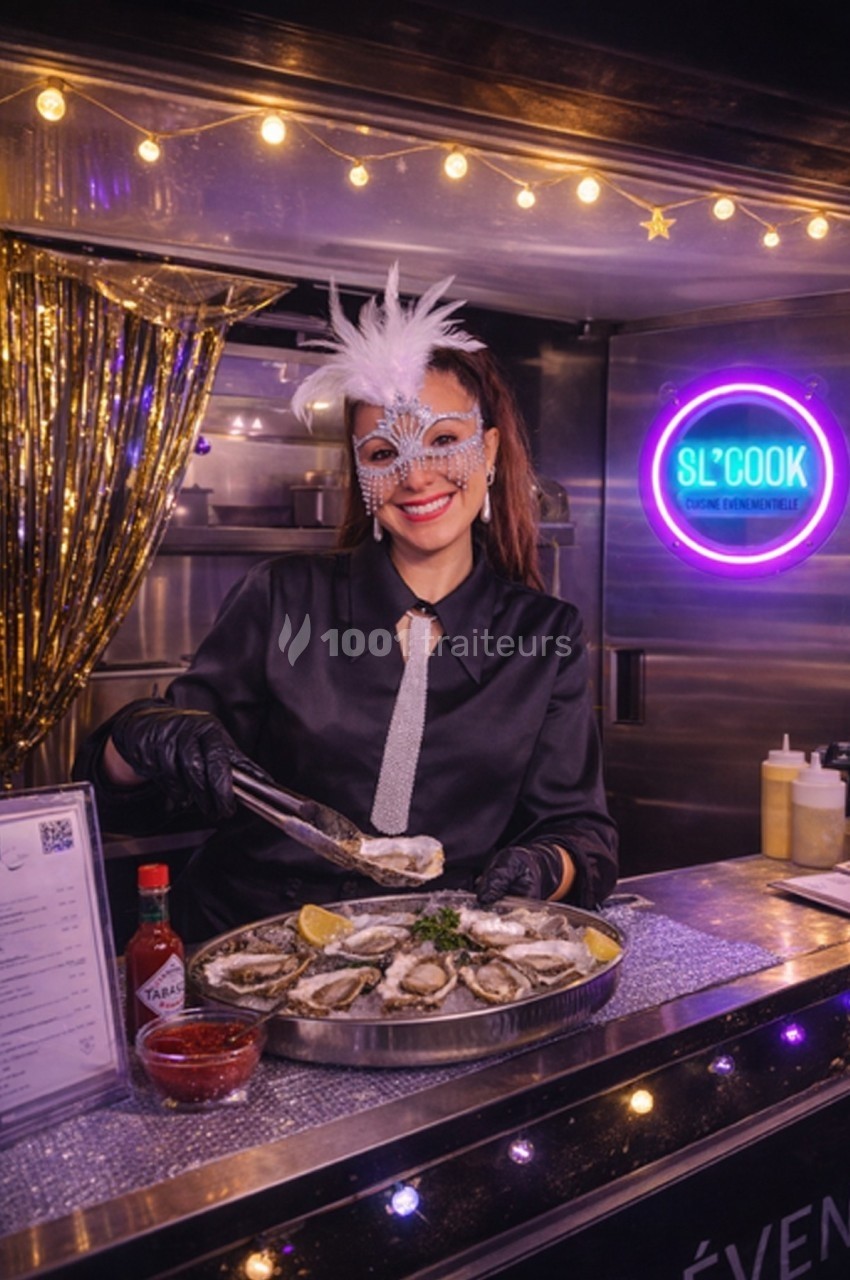 Femme masquée servant des huîtres sur un stand décoré avec des lumières et des éléments festifs.