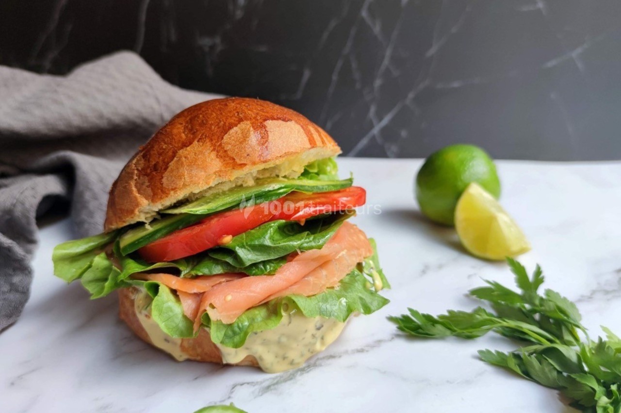 Burger au saumon fumé avec laitue, tomate et sauce, posé sur une table en marbre avec citron vert et herbes en arrière-plan.