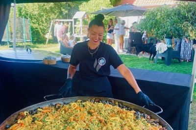 Une cuisinière prépare une grande paella garnie de fruits de mer, de poulet et de légumes lors d'un événement en plein air.