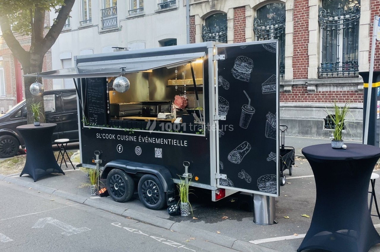 Food truck noir stationné dans une rue, avec des tables hautes et des illustrations de plats sur sa façade.