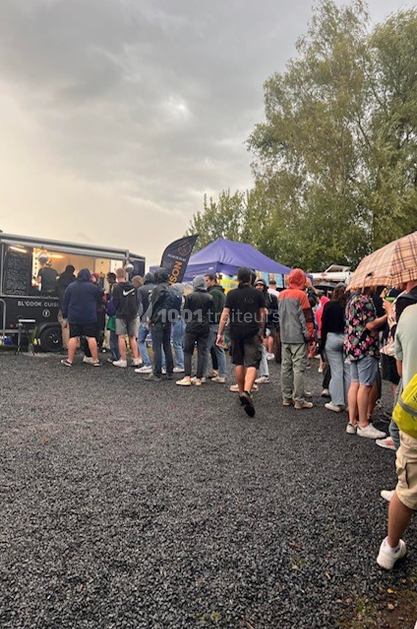 Une file de personnes attendant devant des stands alimentaires et un food truck sous un ciel nuageux.