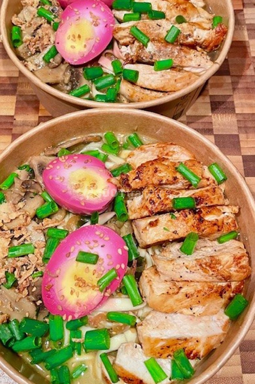 Deux bols de ramen garnis de poulet grillé, œufs marinés roses, champignons et ciboulette sur une table en bois.