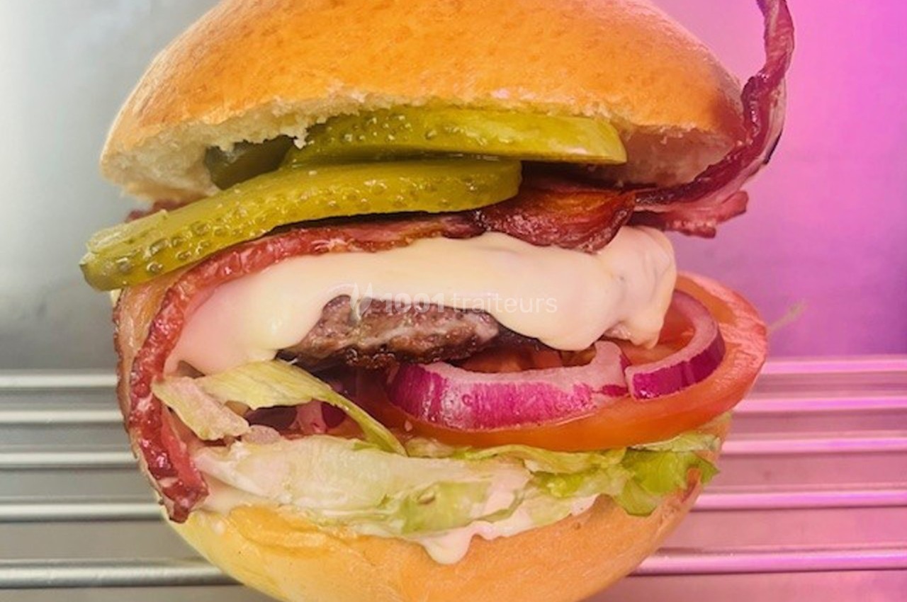 Burger garni de bacon, fromage fondu, cornichons, oignons rouges, tomate et laitue sur une grille métallique.