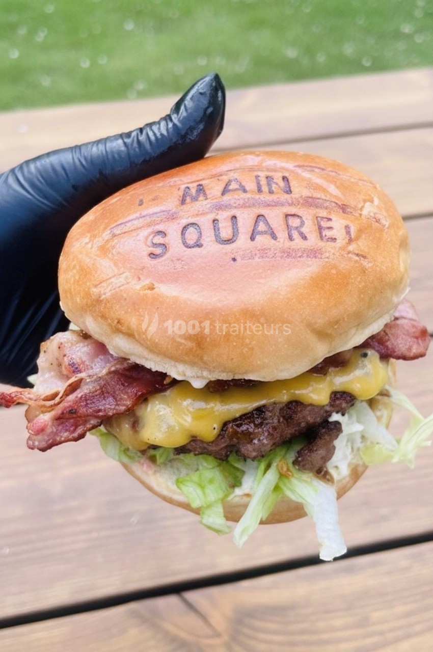 Burger garni de bacon, fromage fondu, laitue et steak haché, tenu par une main gantée sur une table en bois.