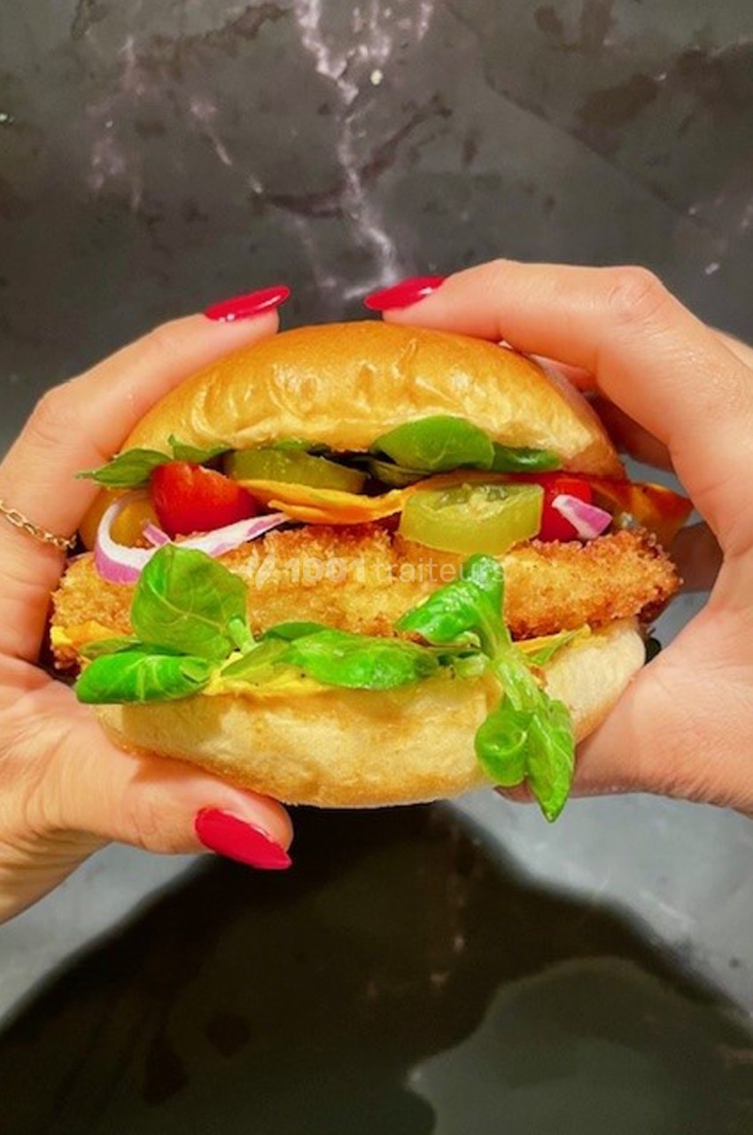 Deux mains tiennent un burger garni de légumes, d'oignons rouges, de cornichons et d'un ingrédient pané.